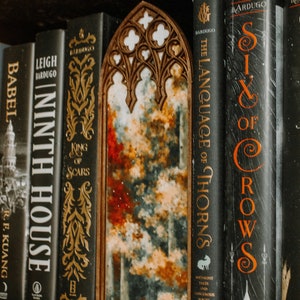 Op de afbeelding: Een houten spiegel met een boogvormige bovenkant, geplaatst tussen boeken. Het spiegeloppervlak toont een kleurrijk, abstract ontwerp met rode, blauwe en gele tinten. De boeken hebben donkere covers met gouden letters.