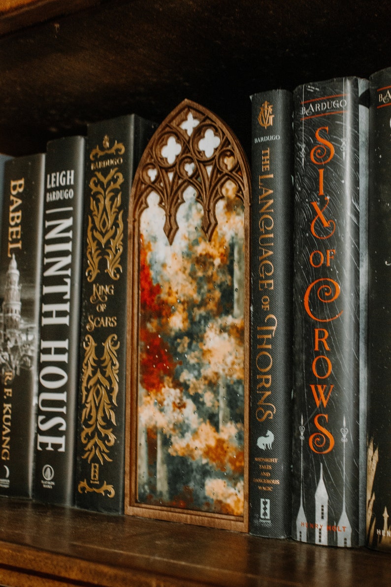 Op de afbeelding: Een houten spiegel met een boogvormige bovenkant, geplaatst tussen boeken. Het spiegeloppervlak toont een kleurrijk, abstract ontwerp met rode, blauwe en gele tinten. De boeken hebben donkere covers met gouden letters.