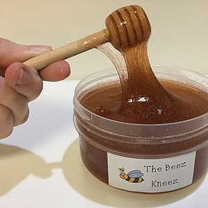 May include: A clear plastic container filled with brown honey-colored slime. A wooden honey dipper is being used to stir the slime. The container has a label that reads "The Beez Kneez" with a cartoon bee illustration.