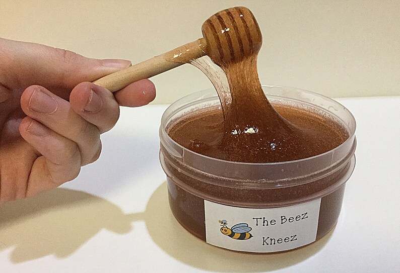 May include: A clear plastic container filled with brown honey-colored slime. A wooden honey dipper is being used to stir the slime. The container has a label that reads "The Beez Kneez" with a cartoon bee illustration.