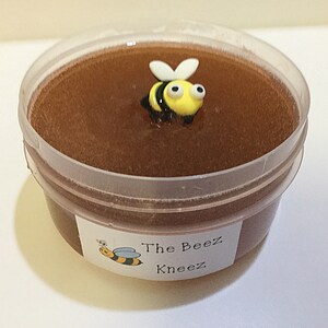 May include: A clear plastic container filled with brown honey slime with a yellow and black bee charm on top. The container has a white label that reads "The Beez Kneez".