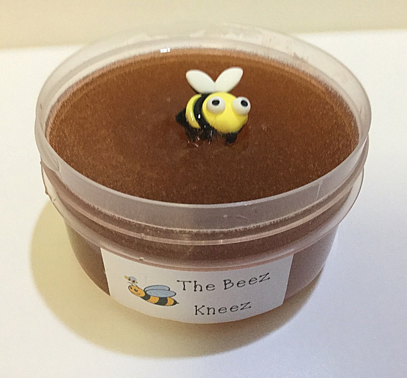 May include: A clear plastic container filled with brown honey slime with a yellow and black bee charm on top. The container has a white label that reads "The Beez Kneez".