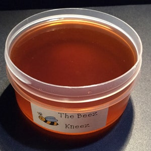 May include: A clear plastic container filled with honey. The container has a label that reads "The Beez Kneez" with a cartoon bee illustration.