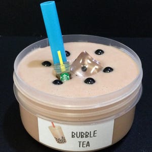 May include: A clear plastic container filled with brown slime, resembling bubble tea. The slime has black boba pearls and a clear ice cube. A blue straw is sticking out of the slime, with a yellow stripe on it.