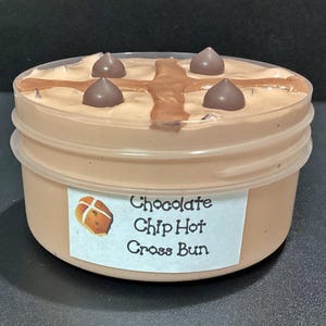 May include: A clear plastic container filled with a brown chocolate chip hot cross bun-flavoured slime. The slime is topped with chocolate chips and a cross made of chocolate.