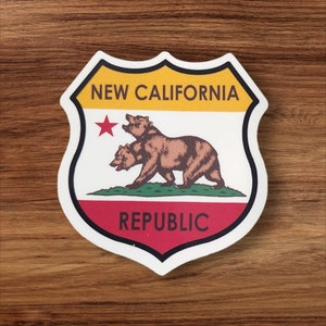 May include: A white sticker with a black outline in the shape of a shield. The sticker features a yellow, red, and green color scheme with a red star and a brown bear. The text "NEW CALIFORNIA REPUBLIC" is printed in black on the sticker.
