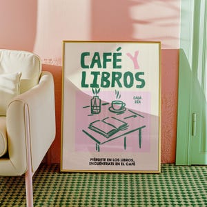May include: A gold framed poster with a pink background and green text that reads "CAFÉ Y LIBROS" with the words "Cada Día" in smaller text below. The poster features a simple illustration of a cup of coffee, a book, and a vase with a flower.