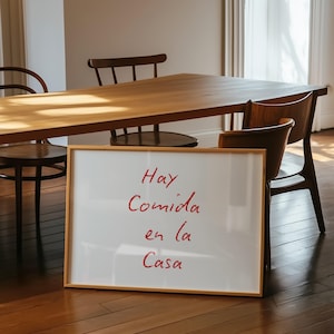 May include: A white framed sign with the words "Hay Comida en la Casa" written in red cursive on a white background. The sign is sitting on a wooden floor in front of a wooden table and chairs.