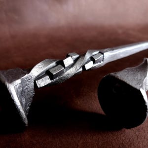 Op de afbeelding: Twee metalen haardgereedschappen met gedraaide en vierkante ontwerpen. De gereedschappen zijn gemaakt van donkergrijs metaal en hebben een rustieke uitstraling.