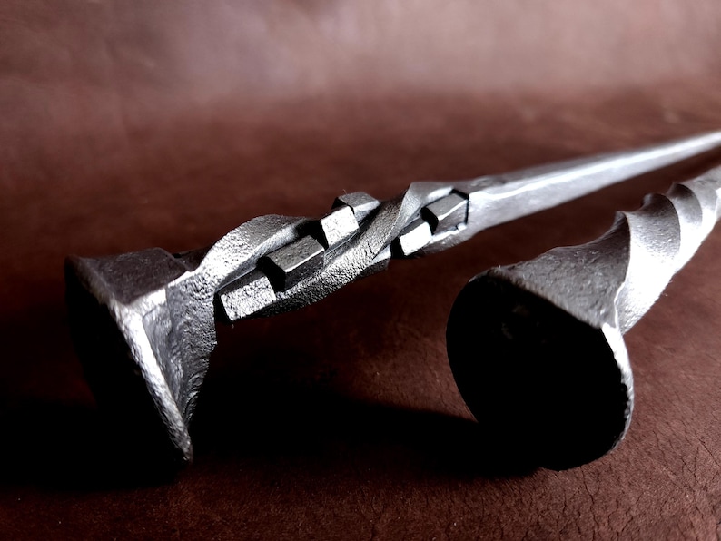 Op de afbeelding: Twee metalen haardgereedschappen met gedraaide en vierkante ontwerpen. De gereedschappen zijn gemaakt van donkergrijs metaal en hebben een rustieke uitstraling.