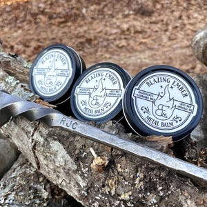 Op de afbeelding: Drie kleine zilveren blikjes met metalen balsem, gelabeld "Blazing Ember" met de ingredi&euml;nten vermeld als "Carnauba wax, bijenwas, minerale olie". De blikjes staan op een stuk hout met de gegraveerde initialen "RJC".