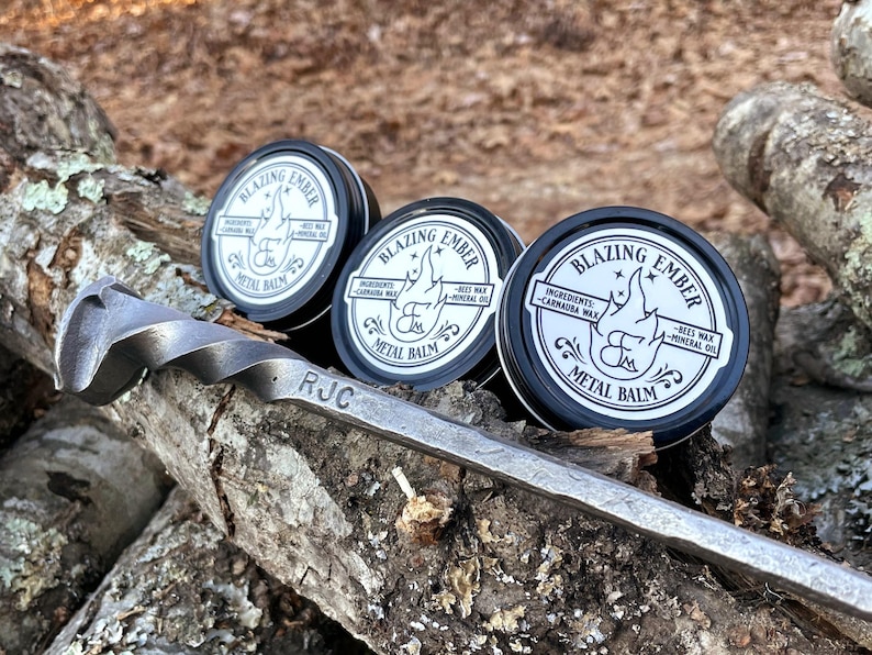Op de afbeelding: Drie kleine zilveren blikjes met metalen balsem, gelabeld "Blazing Ember" met de ingredi&euml;nten vermeld als "Carnauba wax, bijenwas, minerale olie". De blikjes staan op een stuk hout met de gegraveerde initialen "RJC".