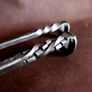 Op de afbeelding: Twee metalen haardgereedschappen met gedraaide handvatten. Het gereedschap bovenaan heeft een eenvoudige draai, terwijl het gereedschap onderaan een meer ingewikkelde draai heeft met vierkante accenten.
