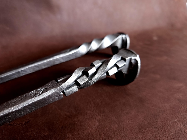 Op de afbeelding: Twee metalen haardgereedschappen met gedraaide handvatten. Het gereedschap bovenaan heeft een eenvoudige draai, terwijl het gereedschap onderaan een meer ingewikkelde draai heeft met vierkante accenten.
