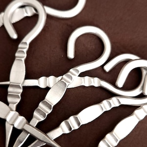 May include: A set of five silver-toned metal hooks with a decorative, textured design. The hooks are arranged in a cluster, with the tips of the hooks pointing in different directions.