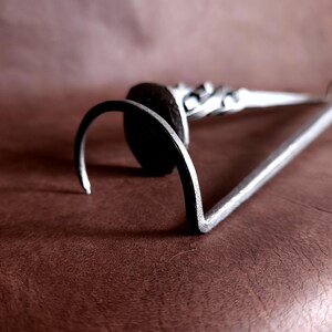 Op de afbeelding: Een zilveren metalen haak met een afgerond uiteinde en een decoratief ontwerp. De haak ligt op een bruin oppervlak.