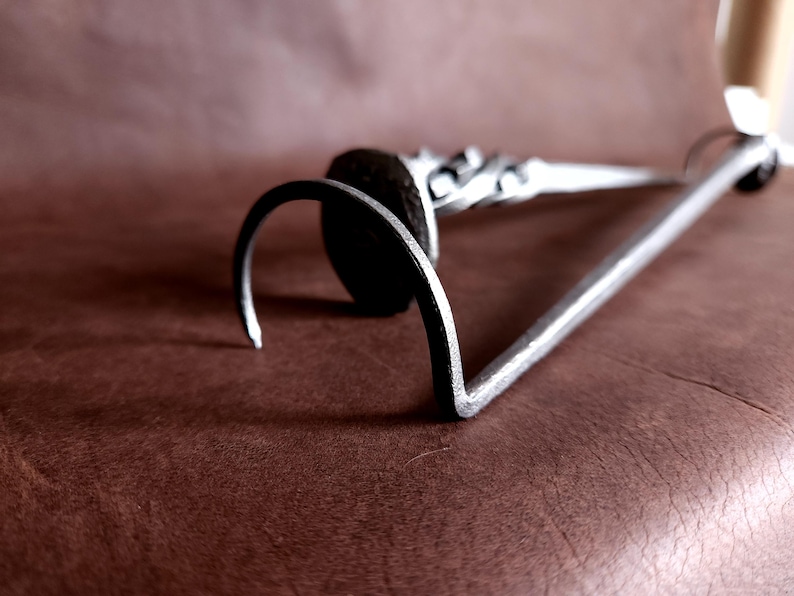 Op de afbeelding: Een zilveren metalen haak met een afgerond uiteinde en een decoratief ontwerp. De haak ligt op een bruin oppervlak.