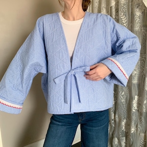 May include: A light blue and white gingham patterned jacket with a tie closure. The jacket has wide sleeves with white trim and red scalloped edges. The jacket is worn over a white shirt and blue jeans.