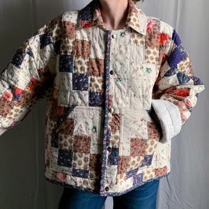 May include: A long-sleeve quilted jacket featuring a patchwork design of various floral and geometric fabric squares in shades of cream, red, blue, and yellow. The jacket has a classic collar, a front button closure with silver snaps, and two pockets.