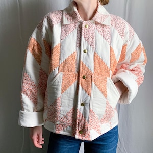 May include: A quilted jacket featuring a chevron pattern with alternating panels of solid cream fabric and floral print fabric in shades of pink and orange. The jacket has a classic collar, long sleeves with folded cuffs, and gold-tone metal snap buttons.
