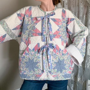 May include: A quilted jacket with a star pattern in shades of blue, pink, and white. The jacket features a round collar, two front pockets, and tie closures. The sleeves have white cuffs, and the jacket is made of a floral patterned fabric.