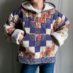 May include: A long-sleeve hooded pullover made from a patchwork quilt featuring a variety of floral and polka dot fabric squares in shades of blue, red, white, and yellow. The garment includes a large front kangaroo pocket and white lining.