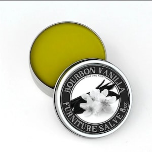 May include: A round tin container with a black and white label that reads "Bourbon Vanilla Furniture Salve 8 oz Wise Owl Paint". The label features a black and white illustration of vanilla beans and white flowers. The tin is open and the container is filled with a yellow salve.