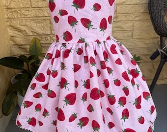May include: A pink sleeveless dress with a strawberry print and white polka dots. The dress has a scalloped hem and a halter neckline.