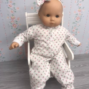 May include: A baby doll wearing a white floral print sleeper and a pink bow headband. The doll is sitting on a white wooden chair.