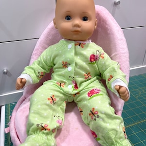 May include: A doll wearing a green and white pajamas with a butterfly and ladybug print sits in a pink plush chair.