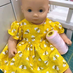 May include: A yellow dress with a bee pattern for a doll. The dress has puffed sleeves and a gathered skirt. The doll is wearing the dress and holding a spool of pink thread.