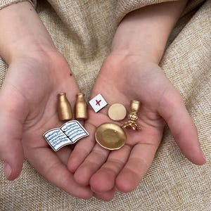May include: A collection of miniature items including two gold-colored bottles, a small square tile with a red cross symbol, a circular gold disc, a gold-colored candlestick, a gold-colored plate, and a flat, open book illustration with blue lines.