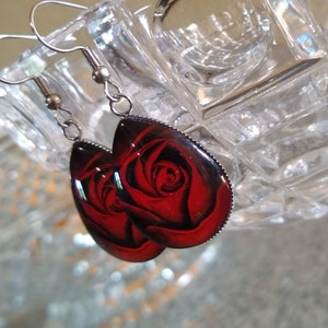 May include: A pair of silver dangle earrings with red rose designs. The earrings are oval shaped and have a silver hook.