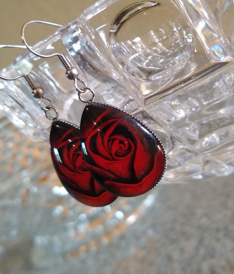 May include: A pair of silver dangle earrings with red rose designs. The earrings are oval shaped and have a silver hook.