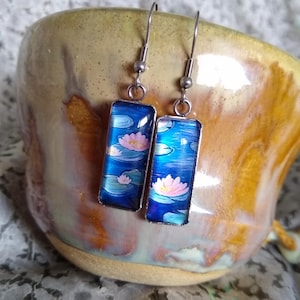 May include: A pair of silver dangle earrings with rectangular blue and white glass charms featuring pink water lilies.