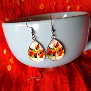 May include: A pair of silver teardrop earrings with a colorful abstract design. The design features geometric shapes in shades of red, orange, yellow, green, and black.