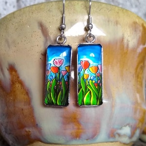 May include: A pair of silver dangle earrings with rectangular glass panels featuring a colorful painting of tulips against a blue sky.