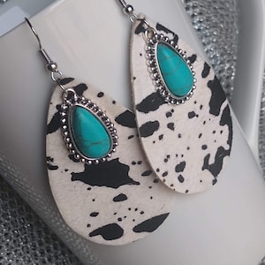 May include: A pair of dangle earrings with a white and black cowhide pattern. Each earring features a turquoise stone set in a silver-toned bezel.