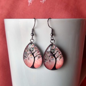 May include: A pair of teardrop-shaped earrings with a tree design. The earrings feature a black tree silhouette with colorful flowers against a pink and white background. The earrings are suspended from silver-colored hooks.