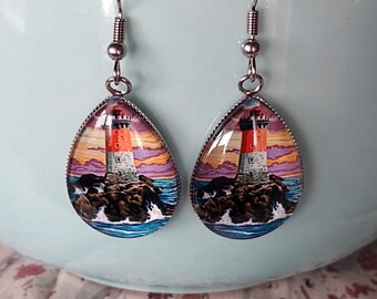 May include: A pair of silver teardrop-shaped earrings with a glass cabochon featuring a lighthouse on a rocky shore with crashing waves and a colorful sunset in the background.