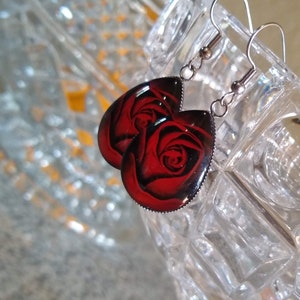 May include: A pair of silver dangle earrings with red rose designs. The earrings are teardrop shaped and have a silver metal frame.