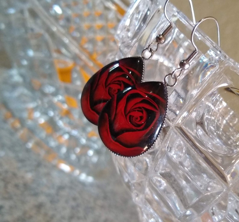 May include: A pair of silver dangle earrings with red rose designs. The earrings are teardrop shaped and have a silver metal frame.