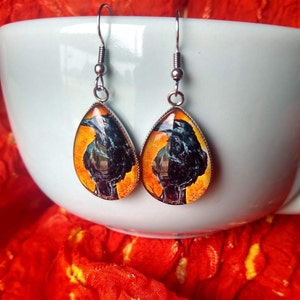 May include: A pair of silver dangle earrings with teardrop-shaped glass pendants featuring a black crow perched on a branch with an orange and yellow background.