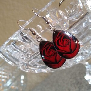 May include: A pair of silver dangle earrings with red rose flower accents. The earrings are teardrop shaped and have a clear glass finish.
