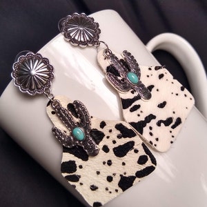 May include: A pair of earrings featuring a cowhide design with black spots and a turquoise stone accent. The earrings are attached to silver-toned metal with a circular design.