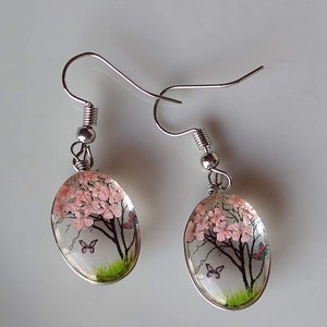 May include: A pair of silver earrings with oval-shaped glass pendants featuring a pink tree with butterflies and green grass.