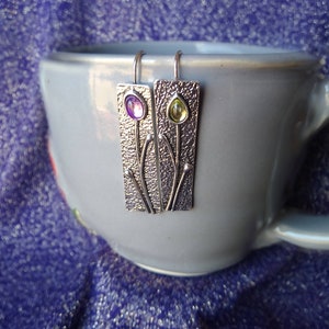 May include: A pair of silver earrings with textured metal and small purple and green gemstones.