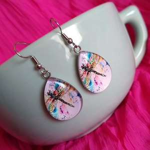May include: A pair of silver dangle earrings with teardrop-shaped glass charms featuring a watercolor dragonfly design on a pink background.