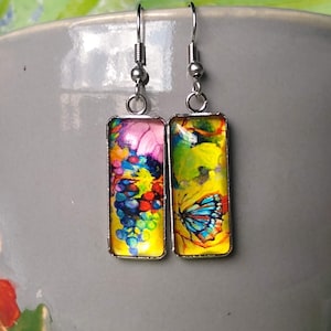 May include: A pair of silver dangle earrings with rectangular glass charms. The charms feature a colorful abstract design with a butterfly and floral elements.