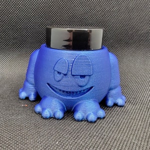 Könnte beinhalten: Eine blaue Keramik-Froschfigur mit einem schwarzen Deckel oben. Der Frosch hat einen schelmischen Gesichtsausdruck.